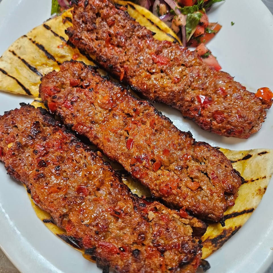 Three grilled Adana kebabs with red peppers on flatbread, served with a side salad.