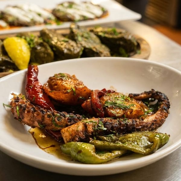 Grilled octopus with red and green peppers, with stuffed grape leaves in the background.