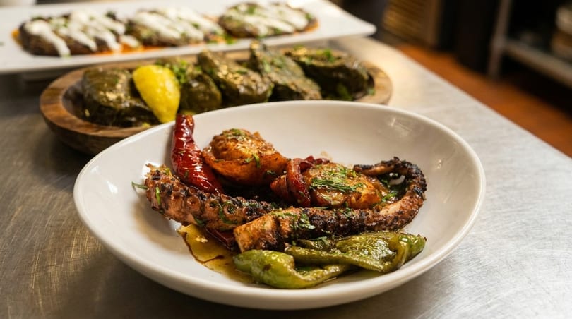 Grilled octopus with red and green peppers, with stuffed grape leaves in the background.