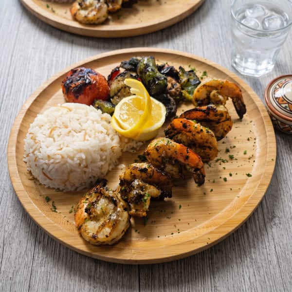 Grilled shrimp, rice, charred tomatoes, and peppers on a bamboo plate with a lemon wedge.